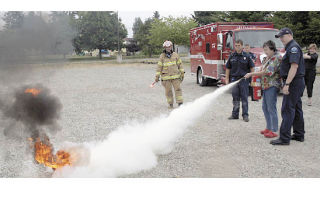 Little Italy owner Carla Lowe is flanked by local firefighters as she practices putting out a small fuel fire with an “ABC” fire extinguisher July 22.